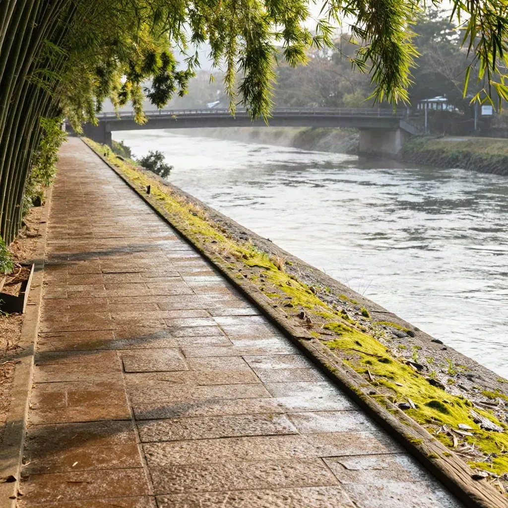 High slope view of Arashiyama path by pale river, bamboo edge, and low bridge in morning mist.